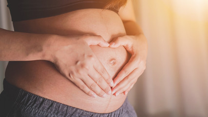 Portrait of young pregnant attractive woman, standing by the window,, Woman holding her hands on her swollen belly shaping a heart,Love concept. Horizontal with copy space.