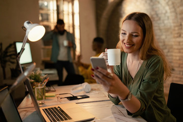 Happy businesswoman text messaging on mobile phone while working late.