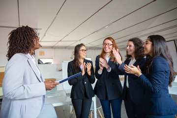 Cheerful coworkers applauding after end of presentation. African American office employee talking while standing with paper documents. Business presentation concept