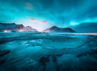 Aurora borealis above the snowy mountain and sandy beach in winter. Northern lights in Lofoten islands, Norway. Starry sky with polar lights. Night landscape with aurora, frozen sea coast, city lights