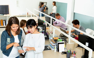 Two smiling girls standing with phone and pointing on screen in modern coworking space with busy colleagues