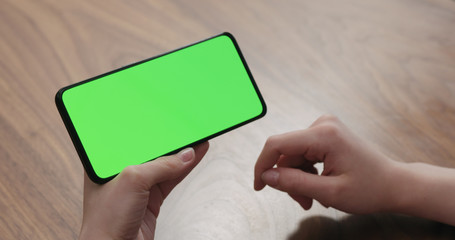 Young woman sitting at a table and using a smartphone with horizontal green screen