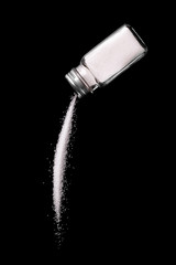 Naklejka na ściany i meble Salt is poured from a salt shaker on a black background. Salt shaker spills salt. A stream of salt.