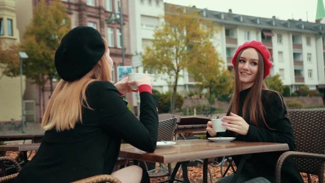 Two beautiful seductive women French in black jackets and red berets sit on the street and drink coffee smiling in a cafe talking at a table. Slow motion, prores