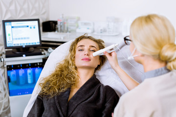 Female beautician does ultrasonic face cleaning and pulse therapy for pretty woman client sitting in a chair in a grey coat in beauty spa clinic