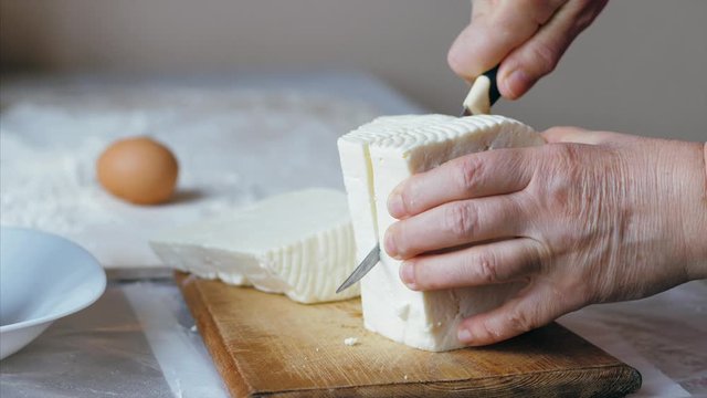 Close-up hands of senior female is cutting a white soft cheese for bakery preparation at domestic kitchen