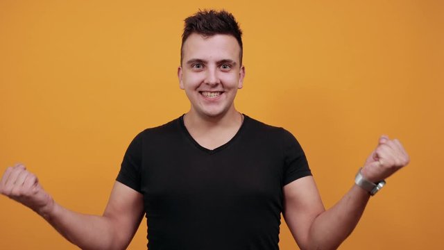 Charming caucasian young man wearing black shirt isolated on orange background in studio doing winner gesture, keeping fists up, smiling. People sincere emotions, lifestyle concept.