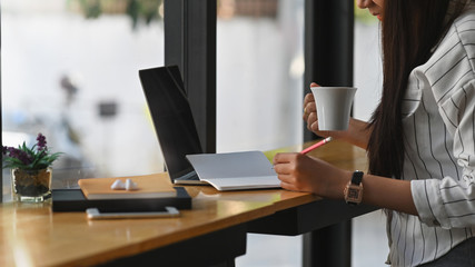 Side shot of young business woman while holding a coffee cup and writing on notebook at the modern office/cafe.