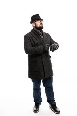 Elegant man with a beard in a coat and hat looks at his watch. Full height. White background. Vertical.