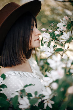 Sensual Portrait Of Beautiful Hipster Woman In Hat Smelling  White Flowers In Spring. Stylish Calm Boho Girl Posing In Blooming Tree With Flowers In Sunny Spring Park. Copy Space