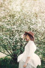Happy boho woman in hat having fun in sunny light near white blooming tree in spring park. Stylish hipster girl enjoying spring and dancing. Atmospheric moment of happiness