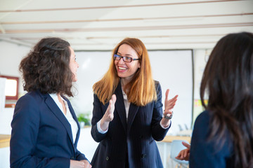 Fototapeta na wymiar Cheerful female colleagues talking. Smiling redhead woman talking and gesturing in office. Communication concept