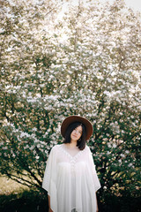 Stylish boho woman in hat posing in blooming tree with white flowers in sunny spring park. Calm portrait of beautiful hipster girl standing in white blooms in spring