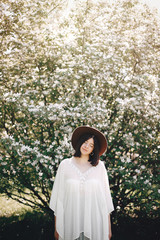 Stylish boho woman in hat posing in blooming tree with white flowers in sunny spring park. Calm portrait of beautiful hipster girl standing in white blooms in spring