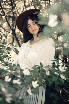 Calm portrait of beautiful hipster girl standing in white blooms in spring. Stylish boho woman in hat posing in blooming tree with white flowers in sunny spring park.