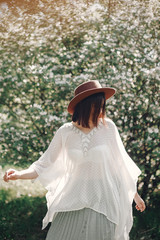 Obraz premium Happy boho woman in hat having fun in sunny light near white blooming tree in spring park. Stylish hipster girl enjoying spring and dancing. Atmospheric moment of happiness