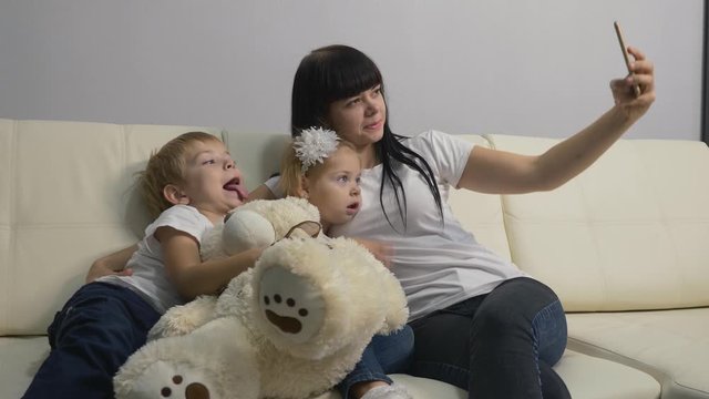 A young mother and small blond children take selfies on the white sofa in the living room. A happy family has fun, uses modern technology in their lives.