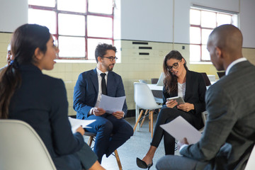 Cheerful managers sitting in chairs with documents and tablet. Confident smiling office employees talking. Teambuilding concept