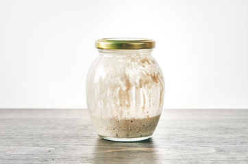 Homemade sourdough starter for bread in a glass jar with closed lid on a table against white background. Made of whole wheat flour. Bubbling fermentation, messy streaks inside container