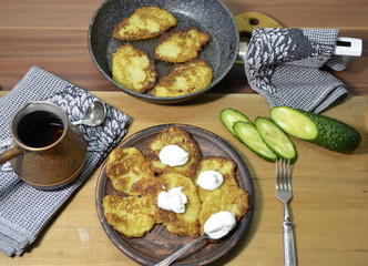 Home kitchen. On the salt there is a frying pan and a plate with appetizing, tasty hot potato pancakes, a Turk with coffee, sour cream, chopped cucumbers, cutlery.