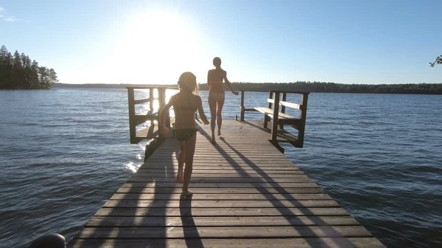 A young slender woman with daughter running on a wooden pier and jumping into the lake after the sauna in summer white night in Finland.