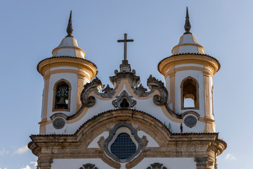 Top facade of the Our Lady of Carmel church in Mariana, Minas Gerais, Brazil, with two round bell towers and cross in the baroque style typical design of architect and sculptor Aleijandinho