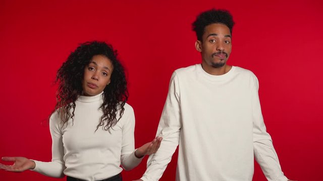 Young african unsure couple shrugs theirs arms, makes gesture of I don't know, care, can't help anything on red background.