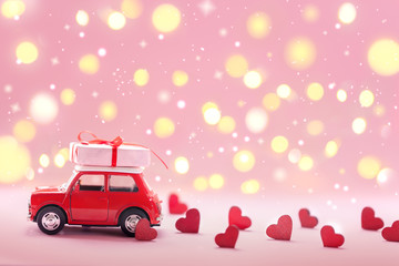 Red toy car delivering carrying on roof gift box with small red hearts on pink background. Valentine's day concept.