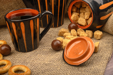 Chunks of brown sugar, a sugar bowl, a mug of tea, and ceramic dishes against a background of coarse homespun cloth. Close up.
