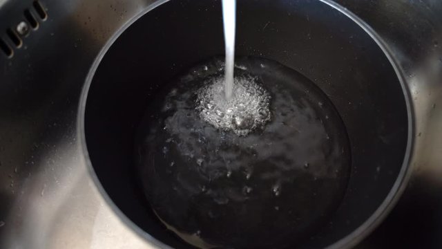 Stainless kitchen pan filled with clean water from the tap. Cooking pan lying in the sink and pure water flowing in it. Water votex and bubbles on the surface, stream of water from the tap, flowing