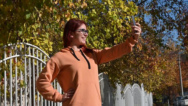 Beautiful red-haired woman in glasses makes selfie on a mobile. A happy girl in an ocher sweatshirt is photographed by a hedge on the phone on a warm autumn day. Leaf fall.