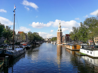 View of Amsterdam canal and Montelbaanstoren tower on a sunny summer day, showing surrounding trees, boats & blue sky.