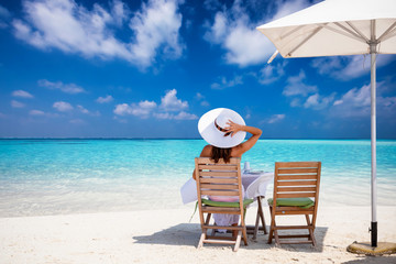 Fototapeta na wymiar Frau in weißem Sommerkleid und mit Hut sitzt beim Mittagessen an einem tropischen Traumstrand mit türkisem Meer und blauem Himmel