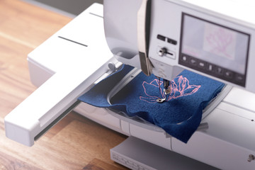 oblique view on modern embroidery machine stitching light pink magnolia graphic on classic blue...