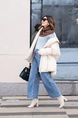 European model girl in a beige oversized down jacket, knitted sweater, flared jeans with a handbag and glasses is walking down the street. Life style