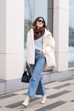 European model girl in a beige oversized down jacket, knitted sweater, flared jeans with a handbag and glasses, walks near the office building. Life style