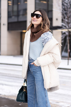 Caucasian model girl in a beige oversized down jacket, knitted sweater, flared jeans with a handbag and glasses, walks in the winter near the office building. Life style
