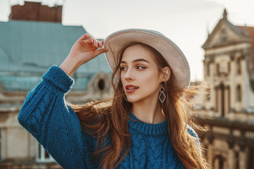 Outdoor close up fashion portrait of young happy brunette woman with long natural hair. Model wearing blue sweater, stylish white hat, trendy earrings, posing at sunset, in European city. Copy space