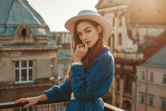 Young elegant fashionable brunette woman, model wearing trendy blue knitted sweater, stylish white hat, wrist watch, earrings, ring, posing at sunset, in European city. Copy, empty space for text