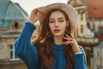 Outdoor close up fashion portrait of young elegant fashionable brunette woman, model wearing trendy blue sweater, stylish white hat, wrist watch, earrings, ring, posing at sunset, in European city - obrazy, fototapety, plakaty