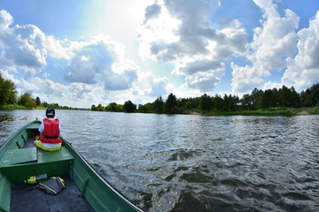 Człowiek w czerwonym kapoku na zielonej łódce płynie rzeką w letni dzień. - obrazy, fototapety, plakaty