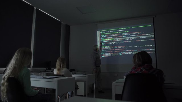 Mature teacher pointing at code projection and explaining data to group of children during computer science lesson in dark classroom