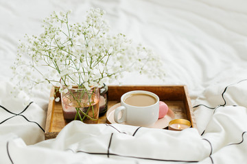Wooden tray of coffee and candles on bed. White bedding sheets with striped blanket and pillow. Breakfast in bed. Hygge concept.