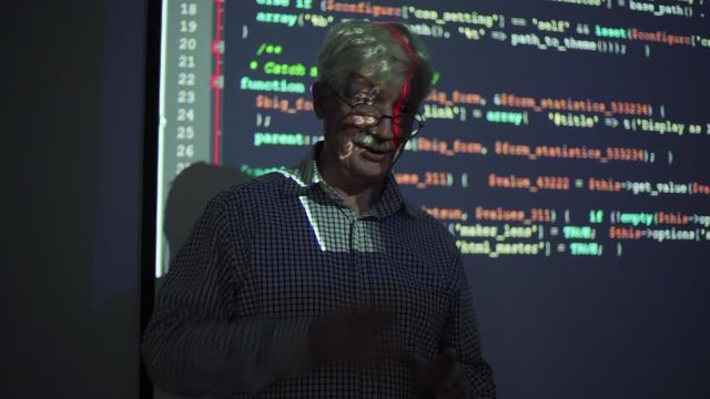 Elderly teacher gesticulating and talking to audience while standing near whiteboard with code projection during programming lesson in dark classroom