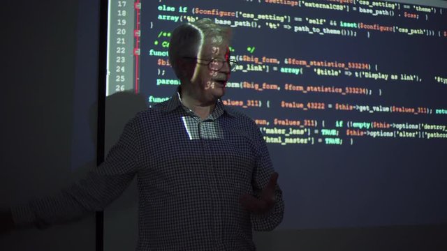 Smiling senior teacher with moustache gesticulating and smiling while explaining code during computer science presentation in dark classroom in modern school