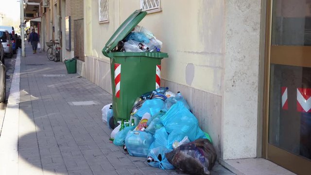 Green plastic container is overfilled with trash and organic waste on the street of Ladispoli, Italy. People throwing out garbage on the ground under the windows of residential building. Trash problem