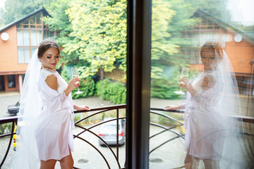 Beautiful bride in a white coat on the balcony drinks champagne from a glass