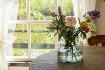 Vase with a mixed bouquet of flowers standing on a wooden table behind a window in the kitchen