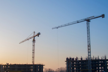 Fototapeta premium Silhouettes of tower cranes constructing a new residential building at a construction site against sunset background. Renovation program, development, concept of the buildings industry.