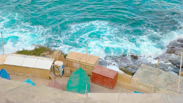 Windy clear blue water crashes on the rocks at the coast of Malta in the Mediterranean near some cargo containers at a construction site. Slowmotion.